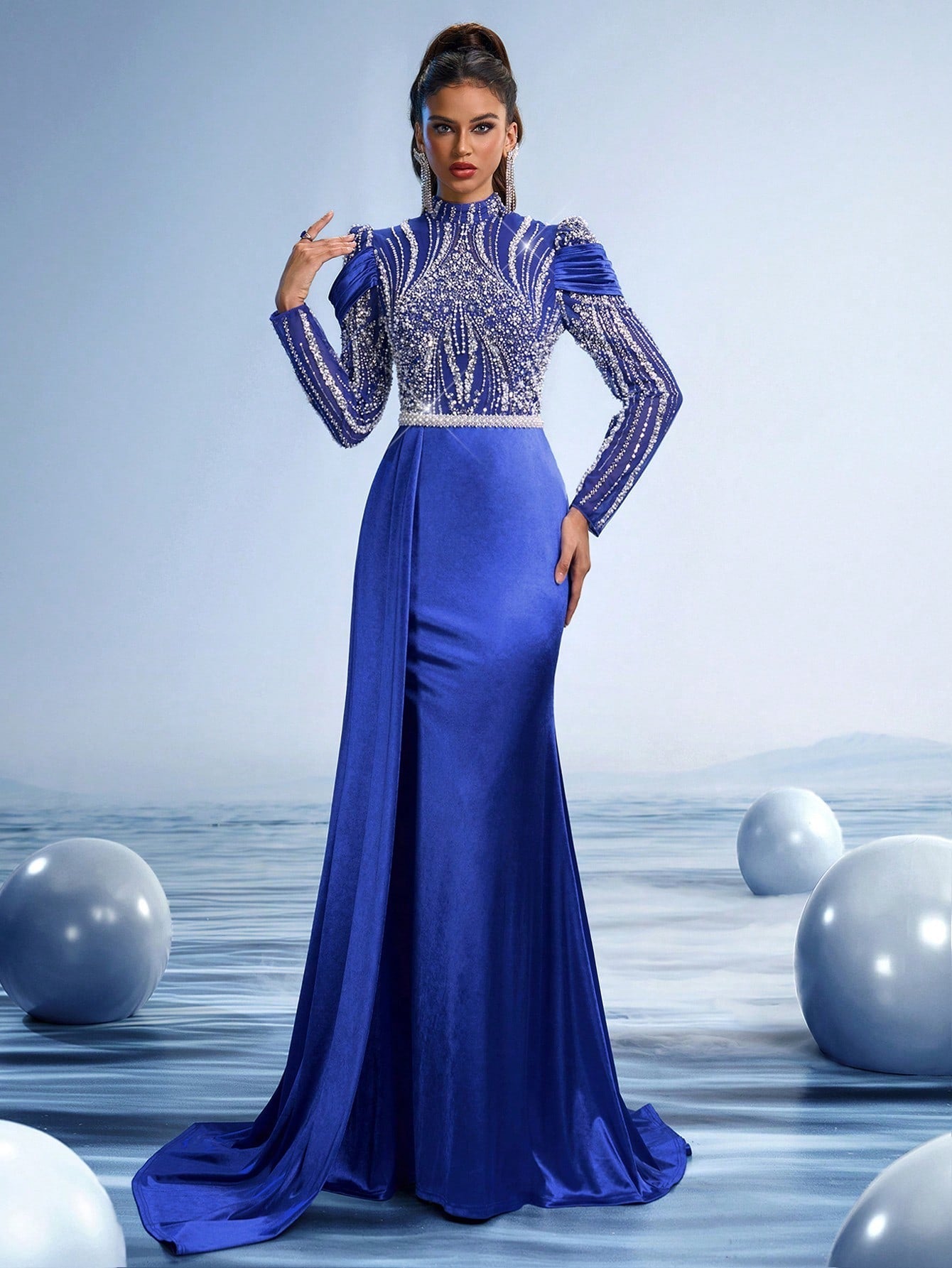 Elegant woman wearing a Sapphire Blue mermaid evening gown with a high neck, intricate silver crystal embroidery on the bodice, long sleeves, and a dramatic floor-length draped cape.