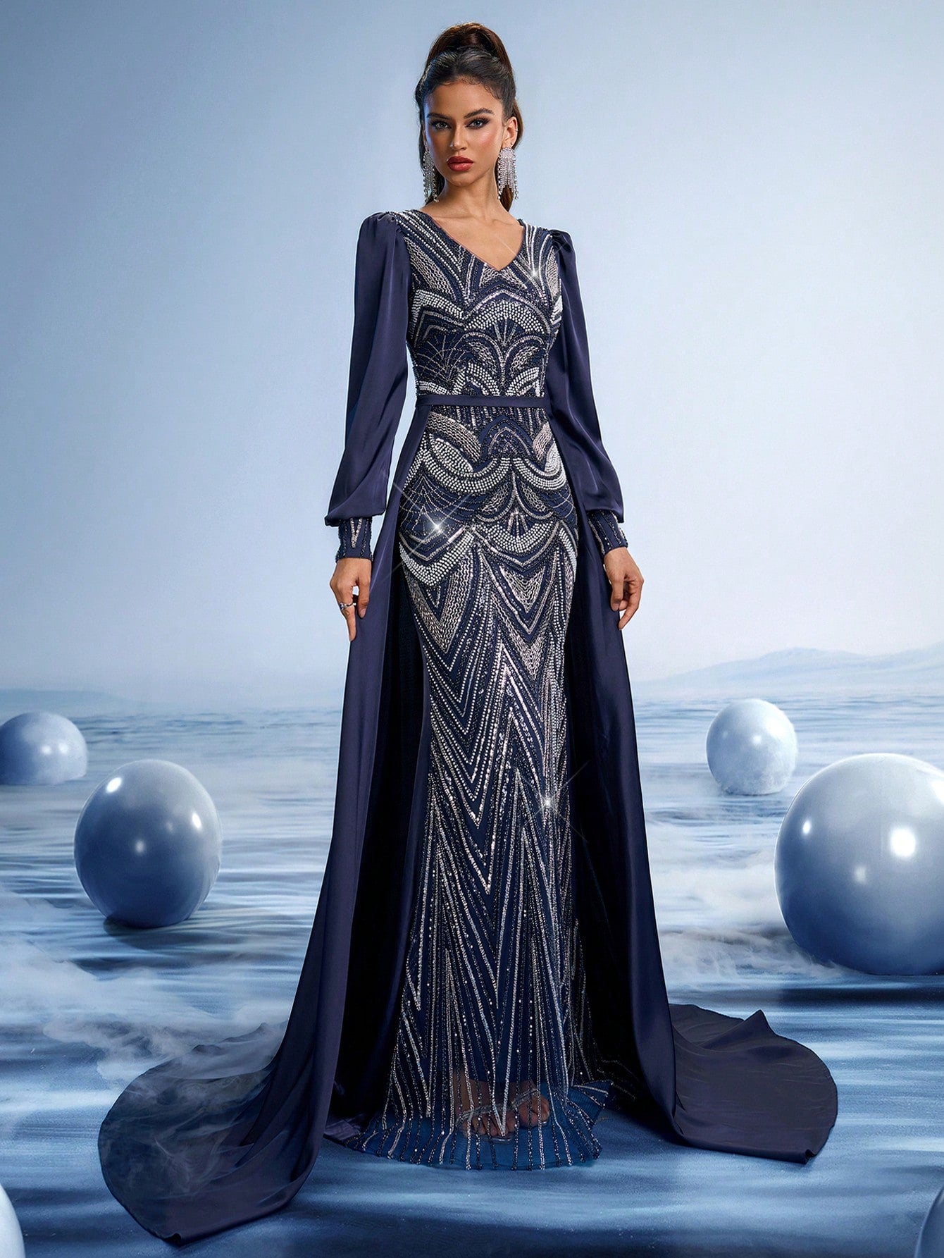 Model wearing a midnight blue evening gown featuring intricate silver metallic embroidery, long bishop sleeves, and a dramatic floor-length cape.