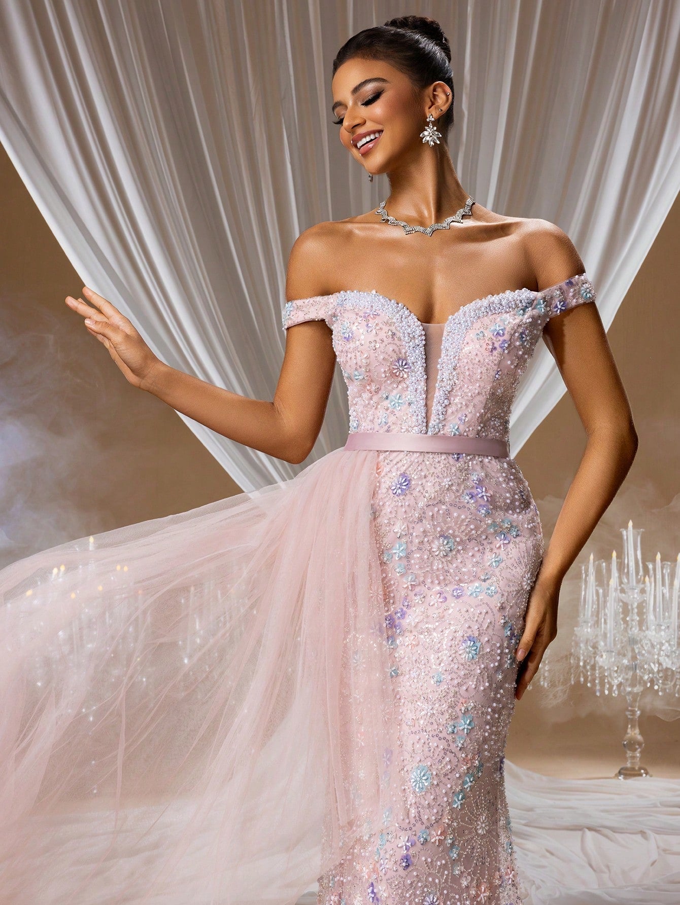 Elegant woman wearing a pink off-the-shoulder evening gown featuring 3D floral lace appliqués, shimmering sequins, and a dramatic layered tulle overskirt for a formal wedding guest or gala event.