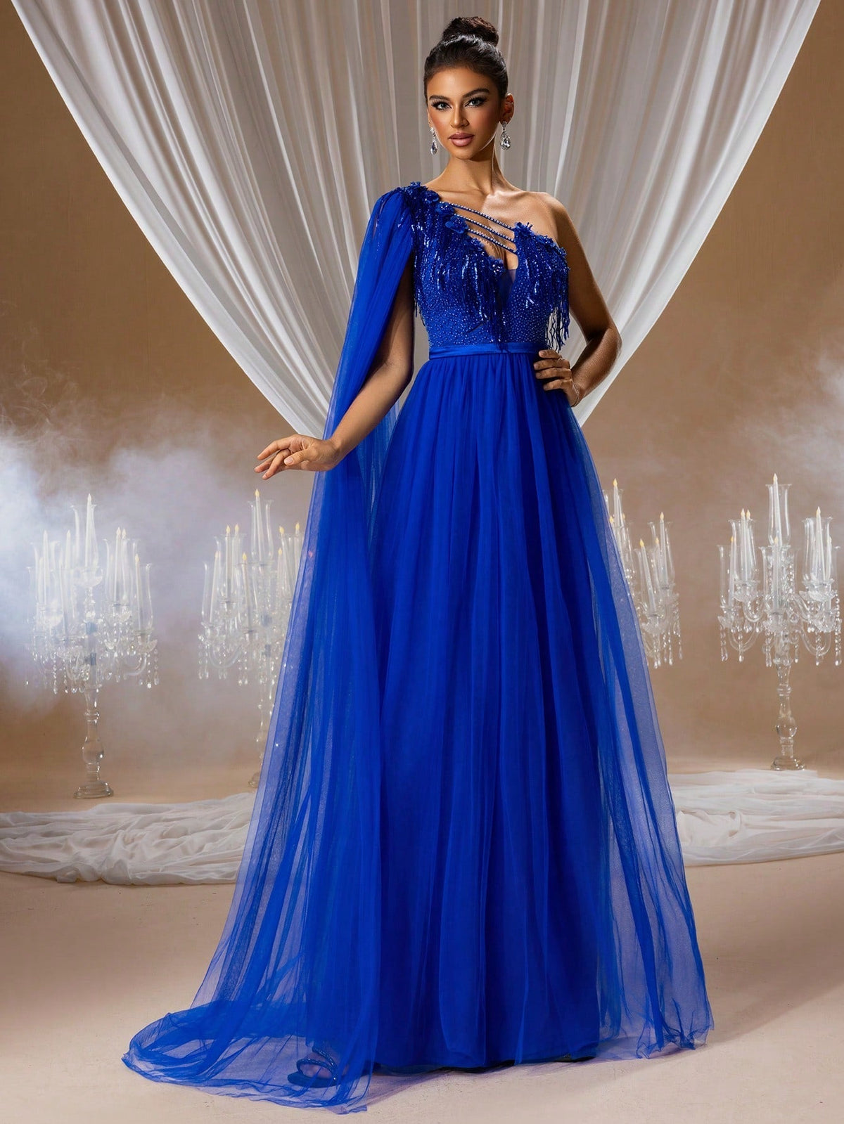 Woman wearing an elegant royal blue one-shoulder evening gown with a long chiffon cape and beaded bodice in a decorated gala setting.